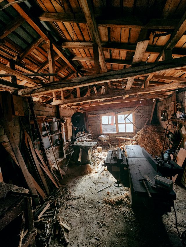 A timber-framed workshop interior with lumber stacked along the walls and a long bench under the window